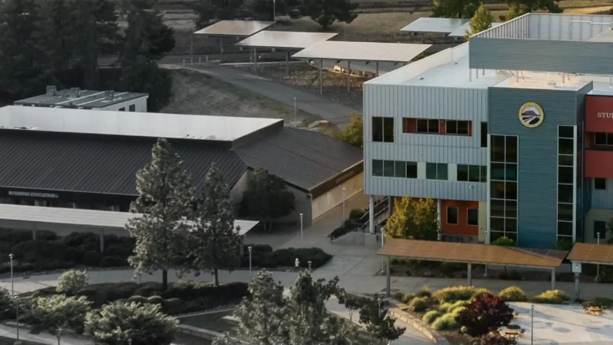 Aerial view of Butte College featuring the modern, multi-story Student Administrative building on the right, rendered in vibrant colors with polished metal siding and large windows. To the left, the older Business Education building appears in muted, darker tones with a low-profile brown roof