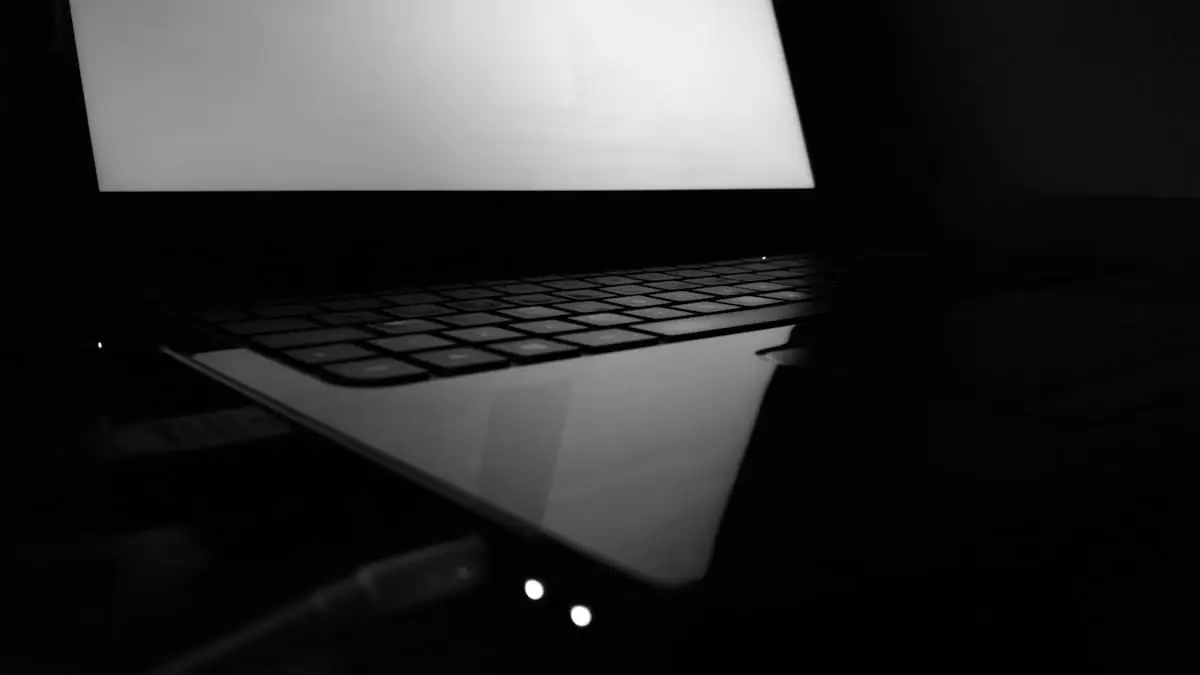A high-contrast, black-and-white photograph of a laptop in a dark room. The screen is glowing bright white, reflecting off the glossy surface of the palm rest. The keyboard and a charging cable plugged into the side are partially visible in the shadows.